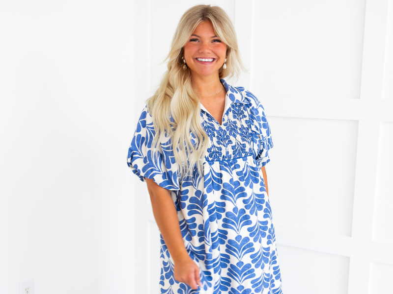 A blonde girl wearing a white and blue patterned dress smiles at the camera